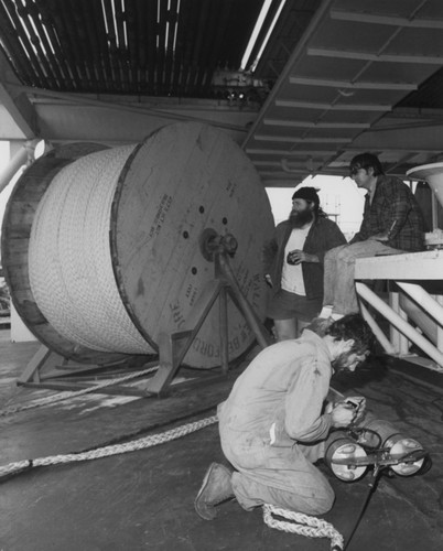 During Leg 65 of the Deep Sea Drilling Project, technicians prepare last-minute adjustments on the downhole instrument package before it is attached to the heavy line seen on the reel after which the array was lowered to the sea floor. 1979