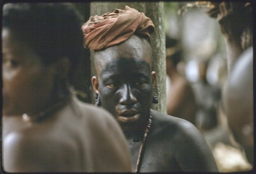 Mortuary ceremony: mourning woman with black paint on face and body, shaved head, wears turtle shell earrings and red shell necklace