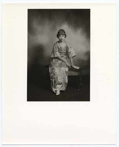Studio portrait of Miss "Flying Phoenix," an actress, dressed in gi pao of the 1920's, seated on a bench /
