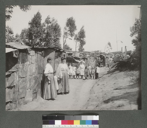 [Women and children refugees posing along a row of makeshift dwellings. Unidentified location.]