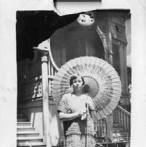 Exterior view of Genevieve Wheeler Cline standing in her wedding dress outside what might be her residence. She is holding a very large decorative umbrella