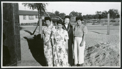 Photograph of four women posing next to a building with two cars in the background at Manzanar