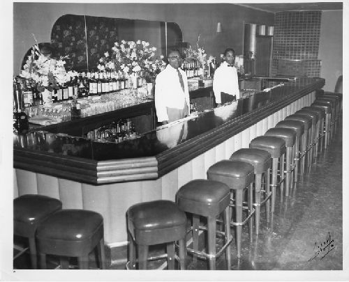 Two male bartenders wearing short white serving coats stand behind bar at Slim Jenkins Restaurant and Bar Oakland, California