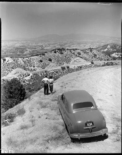 Man and woman standing by their car on a ridge overlooking valley below with mountains in background