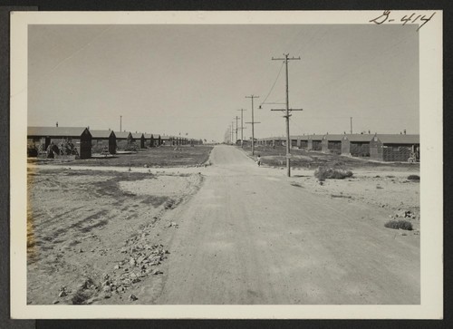 Looking down a street of the Minidoka Relocation Center. A service flag with two stars can be seen in the window of the first room in the barrack at extreme right. Hunt, Idaho
