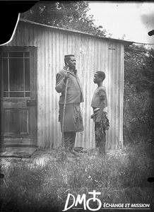 African man and boy standing in front of a corrugated iron building, Antioka, Mozambique, ca. 1901-1915