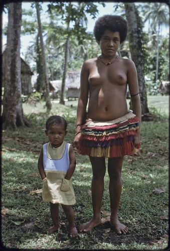 Adolescent girl wearing short fiber skirt stands with young girl in cloth dress, both wear red shell necklaces