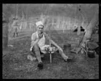 Boy Scout at camp event, wearing an apron and chef's hat, seated at a camp table, circa 1935