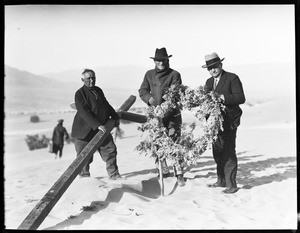 Three men holding a large cross and wreath at a funeral