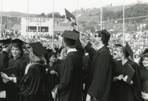 Photograph of students in recessional, a man student holding up his diploma in the Eddie Fields Stadium