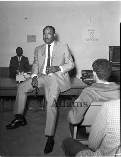 Unidentified man resting against a table in front of an audience, Los Angeles