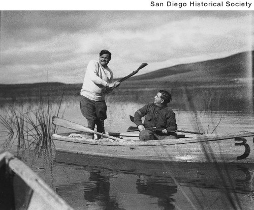 Babe Ruth standing in a rowboat on Sweetwater Lake holding a shotgun like a baseball bat as Linn Platner watches