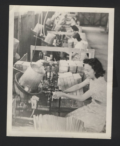Photograph of women in production line, bucket-like machine in front of them, basket slats piled next to worker