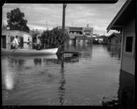 Unidentified men ride boat in flood waters near submerged vehicle and homes, Long Beach, 1930s