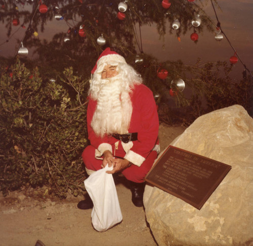 Santa Claus kneeling next to dedicatory plaque at tree lighting ceremony, 1980